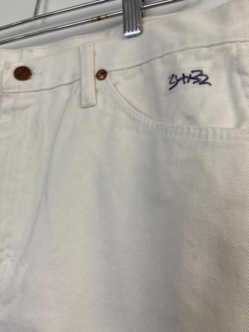Op de afbeelding: Witte spijkerbroek met een paarse handtekening die "3432" op de rechterzak staat.