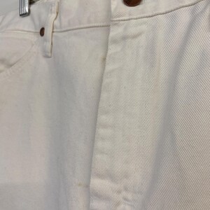 Op de afbeelding: Een paar witte spijkerbroeken met een bruine knoop en een enkele achterzak. De broek hangt aan een hanger.