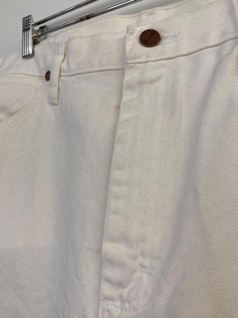 Op de afbeelding: Een paar witte spijkerbroeken met een bruine knoop en een enkele achterzak. De broek hangt aan een hanger.