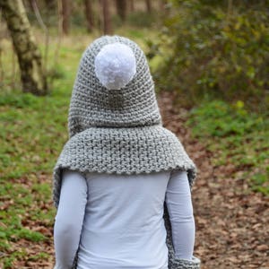 May include: A gray hooded scarf with white pom-poms on the hood and at the ends of the scarf. The scarf is crocheted and has pockets on the sides. The scarf is being worn by a person in a wooded area.