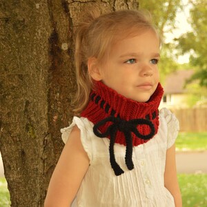 Peut inclure: Une jeune fille portant un col en crochet rouge et noir avec un nœud. Le col est fait avec un fil épais et a un design simple. La fille est debout à côté d'un arbre et regarde de côté. Le texte "Willow Knot Patterns" est visible en bas de l'image.