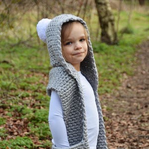 May include: A gray hooded scarf with white pom-poms. The scarf has a pocket on the front and is worn by a child.
