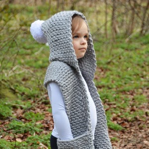 May include: A gray crocheted hooded scarf with white pom-poms. The scarf has a pocket on the front and is worn by a child.