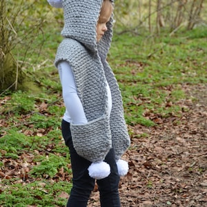 May include: A gray hooded scarf with white pom-poms and pockets. The scarf is crocheted and worn over a white long-sleeve shirt and black pants. The text "Willow Knot Patterns" is written in white on the bottom of the image.