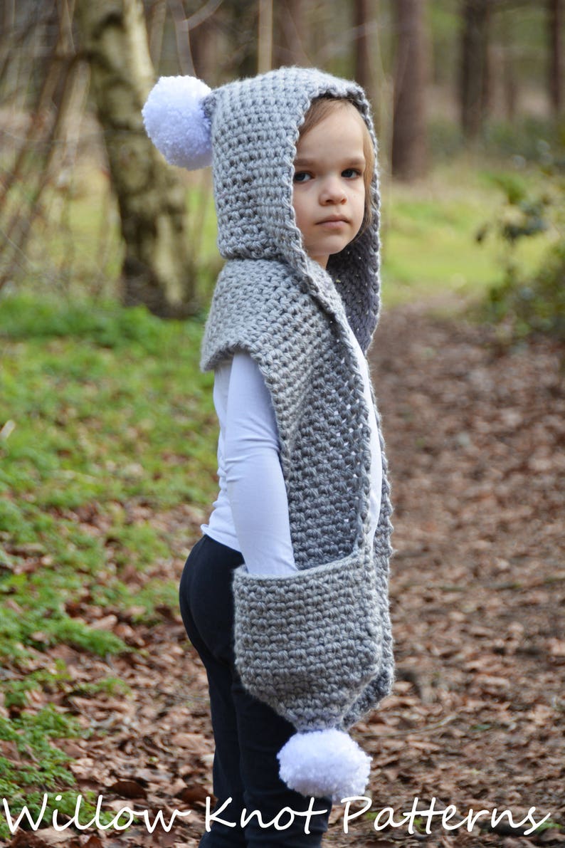 May include: A gray hooded scarf with white pom-poms. The scarf has a pocket on the side and is worn by a child in a forest setting.