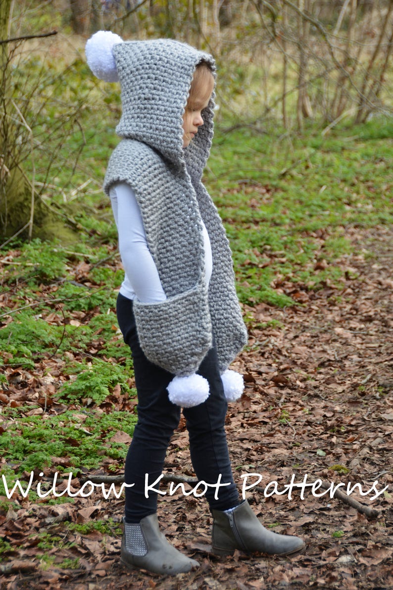 May include: A gray hooded scarf with white pom-poms on the hood and sleeves. The scarf has a pocket on the front. The scarf is worn by a child in a forest setting. Willow Knot Patterns is written on the ground.