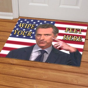 May include: Rectangular doormat featuring an image of a man pointing to his face, with the text "WIPE YOUR FEET HERE" in gold lettering. The background is an American flag design with red and white stripes and a blue field of stars.