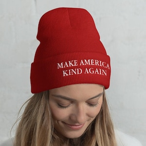 Puede incluir: Gorro de punto rojo con el texto "Make America Kind Again" en blanco.