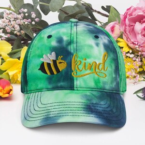 Op de afbeelding: Een groene en blauwe tie-dye baseballpet met een geborduurde bij en het woord "kind" in gouden draad.