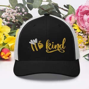 Op de afbeelding: Zwarte en witte truckercap met een gele geborduurde bij en de tekst "be kind" in goud.