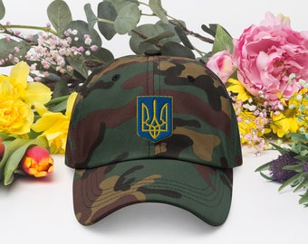 May include: A camouflage baseball cap featuring the Ukrainian coat of arms. The cap has a green, brown, and black pattern. The coat of arms is a gold trident on a blue background. The cap is surrounded by flowers.