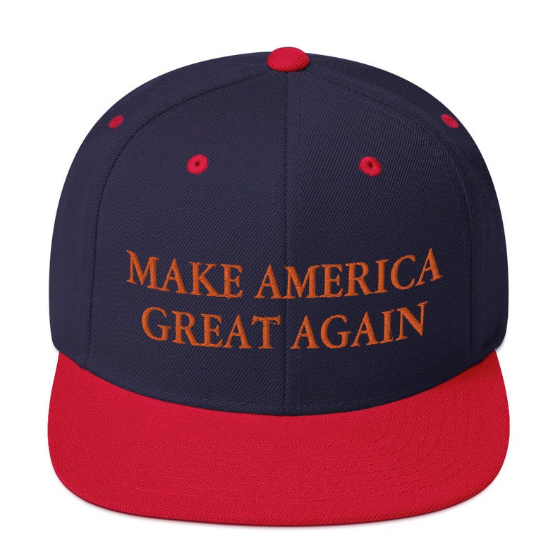 Op de afbeelding: Een marineblauwe en rode baseballpet met de tekst "MAKE AMERICA GREAT AGAIN" geborduurd in oranje.