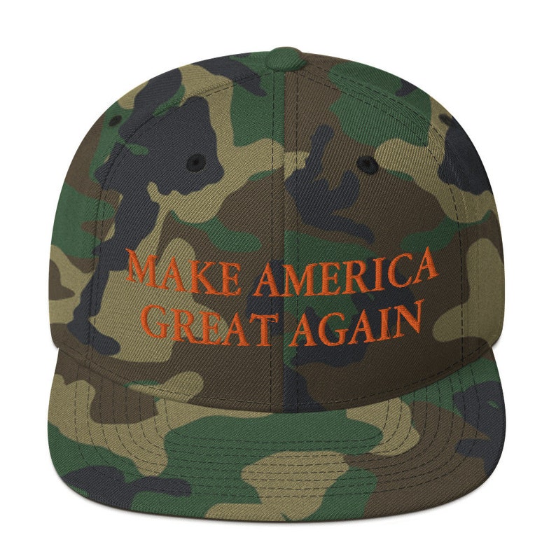 Op de afbeelding: Een camouflagegroene en bruine baseballpet met de tekst "MAKE AMERICA GREAT AGAIN" geborduurd in oranje.