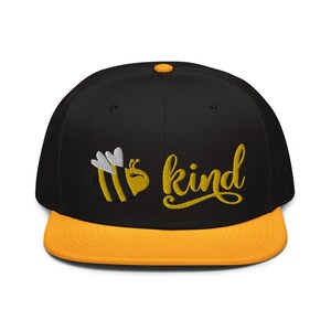 Op de afbeelding: Zwarte en gele baseballpet met een bij en de tekst "be kind" geborduurd op de voorkant. De pet heeft een gele klep.