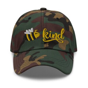Op de afbeelding: Een camouflage baseballpet met een geborduurde gele bij en het woord "kind" in gele schrift.