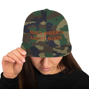 Op de afbeelding: Een camouflage baseballpet met de tekst "MAKE AMERICA GREAT AGAIN" geborduurd in oranje.