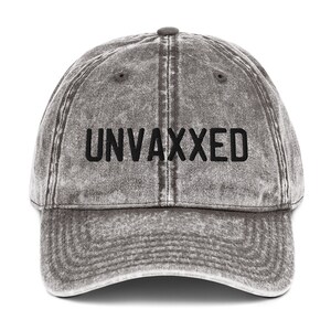 May include: A gray distressed denim baseball cap with black embroidered text that reads "UNVAXXED".