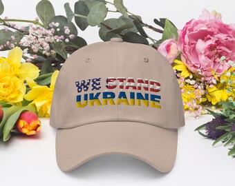 May include: A beige baseball cap with the text "WE STAND WITH UKRAINE" in red, white, and blue, with the letters "UKRAINE" in the colors of the Ukrainian flag.