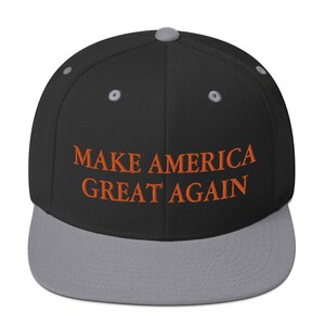 Op de afbeelding: Een zwarte en grijze baseballpet met de tekst "MAKE AMERICA GREAT AGAIN" geborduurd in oranje.