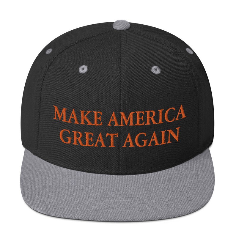 Op de afbeelding: Een zwarte en grijze baseballpet met de tekst "MAKE AMERICA GREAT AGAIN" geborduurd in oranje.