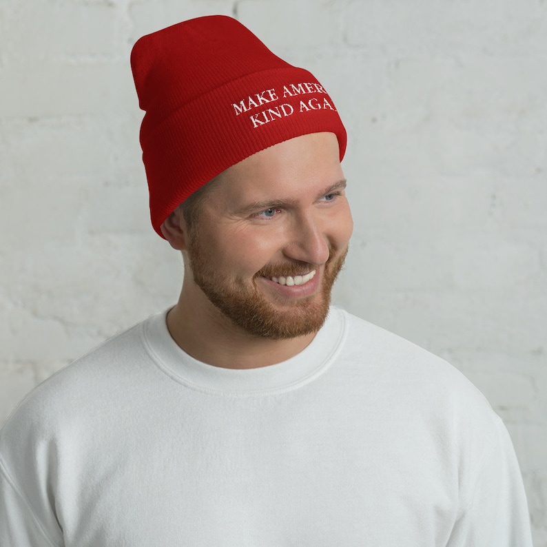 Puede incluir: Un gorro de punto rojo con el texto "MAKE AMERICA KIND AGAIN" en letras blancas.