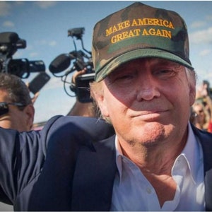 Op de afbeelding: Een man draagt een groene camouflage baseballpet met de tekst "MAKE AMERICA GREAT AGAIN" in gele letters. Hij draagt een wit shirt en een blauw blazer. Hij glimlacht en kijkt naar de camera.