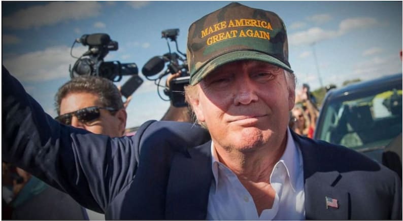 Op de afbeelding: Een man draagt een groene camouflage baseballpet met de tekst "MAKE AMERICA GREAT AGAIN" in gele letters. Hij draagt een wit shirt en een blauw blazer. Hij glimlacht en kijkt naar de camera.
