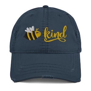 Op de afbeelding: Een marineblauwe baseballcap met een distressed afwerking, met een gele geborduurde bij en het woord "kind". De bij is zwart en geel met witte vleugels. De cap heeft een versleten uitstraling met gerafelde randen.