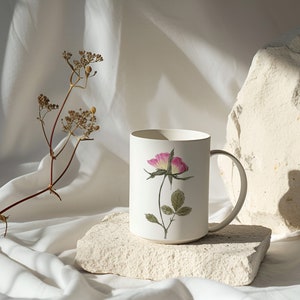 Puede incluir: Taza de cer&aacute;mica blanca con un dise&ntilde;o de rosa rosa sobre fondo blanco. La taza est&aacute; sentada sobre una peque&ntilde;a roca blanca. Un tallo de flor seca est&aacute; en el fondo.