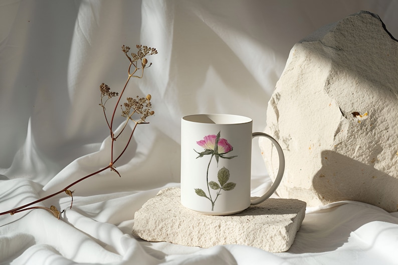 Puede incluir: Taza de cer&aacute;mica blanca con un dise&ntilde;o de rosa rosa sobre fondo blanco. La taza est&aacute; sentada sobre una peque&ntilde;a roca blanca. Un tallo de flor seca est&aacute; en el fondo.