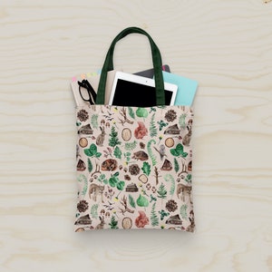 Op de afbeelding: Een beige tote bag met een groene handvat en een bosmotief. Het patroon bevat kleine huisjes, bomen, dieren en bladeren. De tas is gevuld met een tablet, een notitieboekje en een bril.