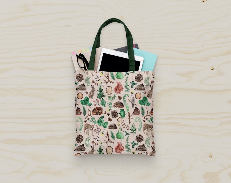 Op de afbeelding: Een beige tote bag met een groene handvat en een bosmotief. Het patroon bevat kleine huisjes, bomen, dieren en bladeren. De tas is gevuld met een tablet, een notitieboekje en een bril.