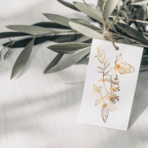 May include: A white gift tag with a gold foil floral design. The tag is tied to a branch of olive leaves.