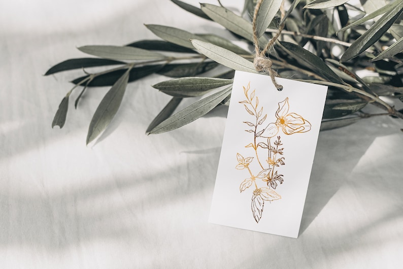 May include: A white gift tag with a gold foil floral design. The tag is tied to a branch of olive leaves.