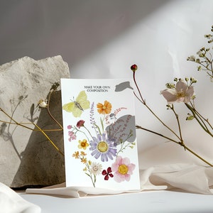 Puede incluir: Una tarjeta blanca con el texto "Make Your Own Composition" y una ilustraci&oacute;n de acuarela de una mariposa y flores. La tarjeta est&aacute; sobre una superficie de tela blanca con flores secas.