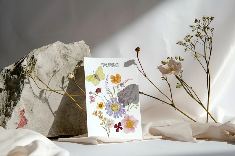 Puede incluir: Una tarjeta blanca con el texto "Make Your Own Composition" y una ilustraci&oacute;n de acuarela de una mariposa y flores. La tarjeta est&aacute; sobre una superficie de tela blanca con flores secas.
