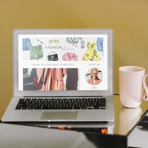 Pode incluir: Um computador port&aacute;til exibindo um site com o texto "FASHION, Stories" e imagens de roupas. O site tamb&eacute;m tem o texto "Trends you don't want to miss out!" e "About Me". Uma caneca rosa com al&ccedil;a de la&ccedil;o est&aacute; em uma mesa de vidro ao lado do laptop.