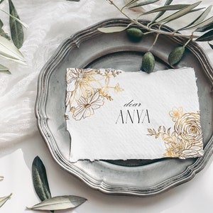 May include: A white card with gold floral illustrations and the text "dear ANYA" on a silver plate with green olive branches.