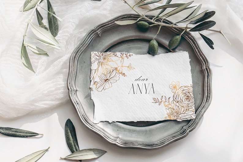 May include: A white card with gold floral illustrations and the text "dear ANYA" on a silver plate with green olive branches.