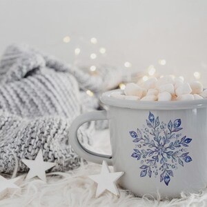 May include: A white enamel mug with a blue snowflake design filled with white marshmallows. The mug is sitting on a white fluffy surface with a grey knitted blanket in the background.