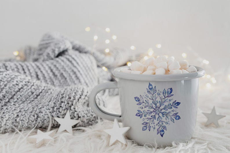 May include: A white enamel mug with a blue snowflake design filled with white marshmallows. The mug is sitting on a white fluffy surface with a grey knitted blanket in the background.