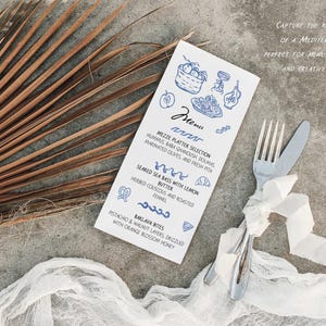 May include: A flat lay featuring a menu with blue illustrations and text, a fork and knife tied with a white ribbon, and a dried palm leaf. The menu lists Mediterranean dishes.