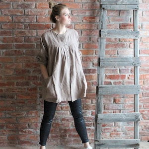 May include: A woman wearing a long, loose-fitting, light brown linen tunic with a button-up front and pockets. She is wearing black leggings and white sneakers.