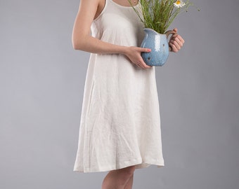 May include: A woman in a white linen dress holds a blue ceramic pitcher filled with a bouquet of blue and white wildflowers.
