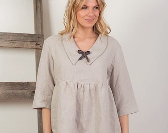 May include: A light beige linen tunic with a wide collar and a black bow tie. The tunic has a gathered waist and 3/4 sleeves.
