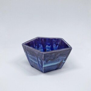 May include: A hexagonal ceramic bowl with a deep blue glaze. The bowl's exterior has a marbled effect with shades of blue and hints of turquoise. The interior is a darker, glossy blue. The bowl is a decorative item.