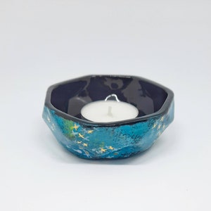 May include: A teal and navy blue ceramic candle holder with a white tea light candle inside. The exterior has a textured, starry design with gold accents. The candle holder has an irregular, hexagonal shape.