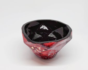 May include: A small, red ceramic bowl with a black interior and gold accents. The bowl has a unique, irregular shape.