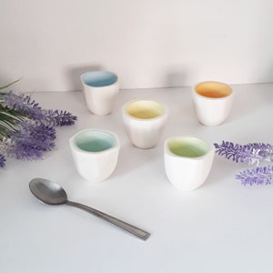 May include: Five small white ceramic cups with different coloured interiors. The cups are arranged in a semi-circle on a white surface. A silver spoon is in the bottom left corner of the image.
