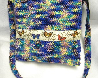 May include: A handmade, crocheted shoulder bag in shades of blue, yellow, and pink. A cream-colored ribbon with butterfly illustrations and the word "Fly" is sewn across the front. The strap is a matching crocheted material.